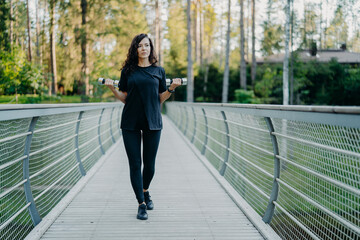 Sporty European woman in black t shirt and leggings, walks across bridge, holds two dumbbells,...