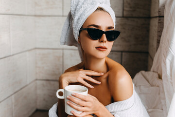 Young woman in white bathrobe, a towel on head and sunglasses with a cup of coffee.
