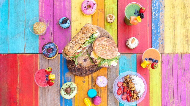 Colorful and tasty drinks and snaks with smoothies, cupcakes, donuts and sandwiches on a colorful wood table - Powered by Adobe