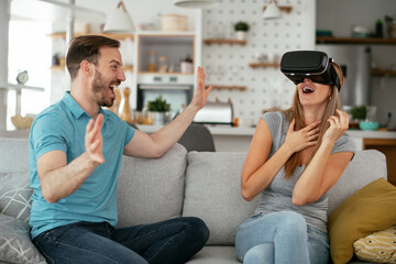 Smiling young woman using VR headset at home on couch. Woman and her husband enjoying virtual reality in apartment.