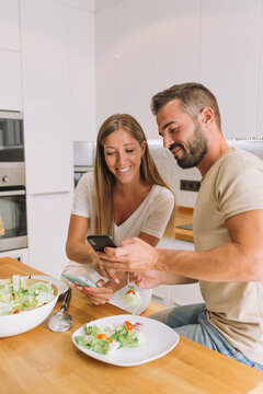 Lovely couple having breakfast and looking photos in mobile phone in the kitchen at home