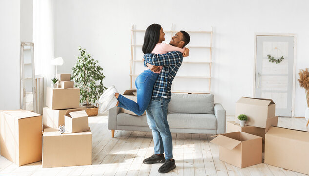 Home relocation concept. Happy black guy hugging and lifting his lovely wife in their new apartment, panorama