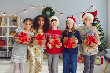 Happy diverse children holding Christmas presents and looking at camera standing in cozy living-room