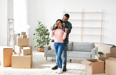 Moving day concept. Millennial black man closing his girlfriend's eyes in their home with cardboard...