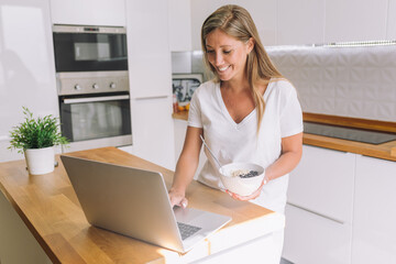 Young blonde woman holding credit card and using laptop computer in home kitchen.	
