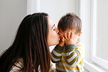 Portrait of young mother kissing her little son in forehead at home. Beautiful female with straight...