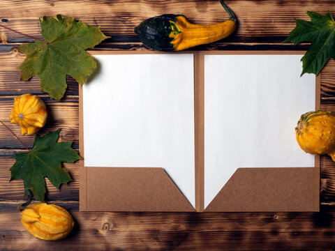 Two white pages for copy space, in a cardboard folder on a burnt wood background with autumn leaves and decorative pumpkins.