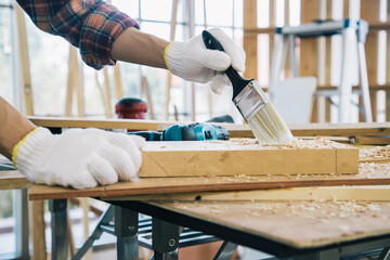 Close up, Carpenter man using soft paint brush clean lumber on wooden table in carpentry office, he is wearing safety gloves for protection to make things safe. Carpentry concept