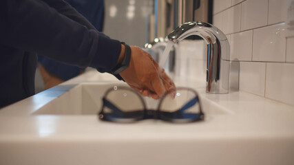 Close up of man putting glasses on top and washing hands in public toilet