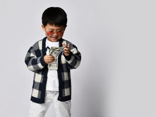 Smiling asian rich kid boy millionaire in modern sunglasses counts dollars cash money he holds in...