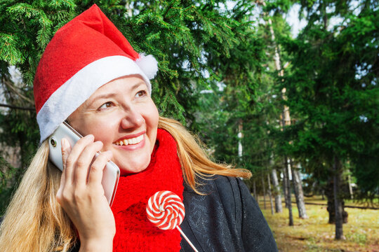 A woman in a Santa Claus hat stands in the park under a Christmas tree, has fun talking on a mobile phone and holds a lollipop in her hand. Christmas and New Year concept