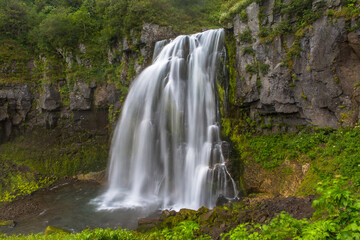Panoramic view of a small waterfall from a cliff overgrown with trees and bushes