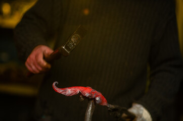 Close-up of a blacksmith's hands with a hammer and a red-hot workpiece in the form of a plant. Work...