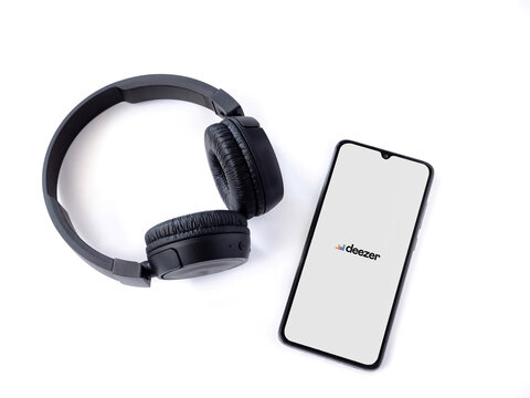 Lod, Israel - July 8, 2020: Black mobile smartphone with Deezer Music Player app launch screen with logo and wireless headphones on a white background. Top view flat lay with copy space.