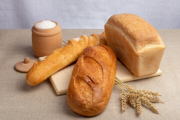 loaf of white bread, baguette and wheat ears on a wooden cutting board and a jar of salt on the...