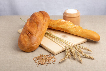 loaf of white bread, baguette and wheat ears on a wooden cutting board and a jar of salt on the...