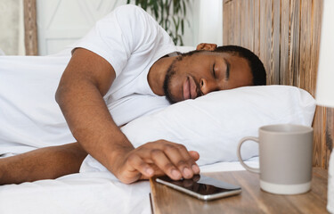 Obraz na płótnie Canvas African Man Sleeping Through Alarm-Clock On Smartphone Lying In Bed