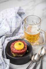 Gourmet Rindertatar Beef Tatar roh aus Filet fettarm, Kapern, Eigelb, Zwiebeln auf Toast Brot und schwarz Teller mit Bier, Löffel, Geschirrtuch und Marmor Hintergrund hell