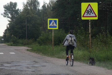 Russian rural woman rides a folding road bike on the side of an asphalt countryside road on a summer evening against the background of green trees and road signs pedestrian crossing, village transport