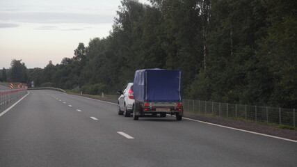 Fototapeta na wymiar Compact passenger car with awning single axle trailer drive on dry asphalted empty suburban highway road on forest background an summer evening, backside view
