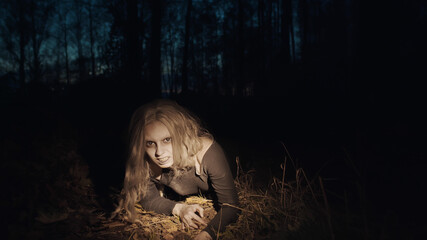 Scary female zombie crawling on ground in night wood illuminated with flashlight