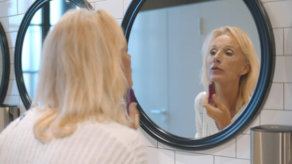 Reflection of attractive middle-aged woman applying lipstick in public restroom