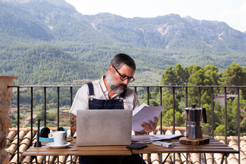 Man with a white beard sitting with a laptop on the terrace in a large window. Man using laptop, shopping or working online from home outdoors. Independent and autonomous