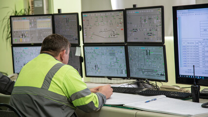 A male engineer, in special clothing, sits against the background of monitors depicting process...