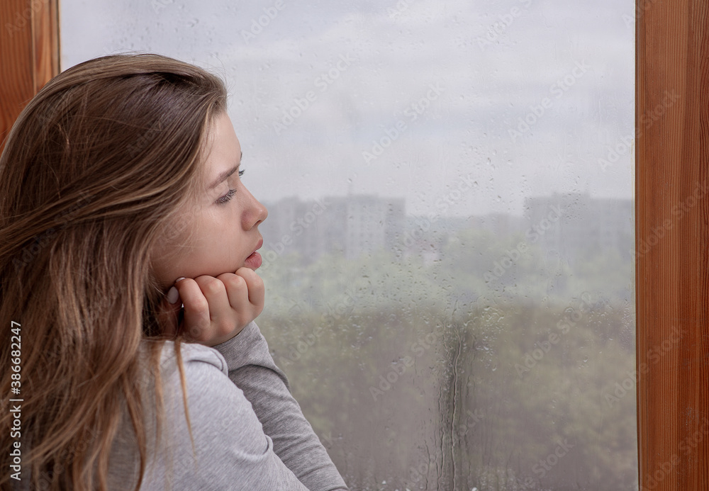 Wall mural sad young woman looking out window, lost in thought, copy space. seasonal depression concept