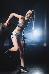 Sportswoman in sportswear and headband exercising with tire during training in gym with smoke