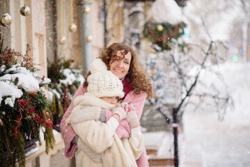 Mom and daughter of European appearance are walking among the Christmas decorations under the falling snow and hugging