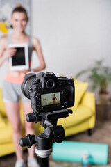 Selective focus of smiling sportswoman with digital tablet standing near digital camera at home