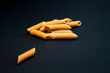 Wholegrain noodles on black background. Penne rigate type, tube shaped macaroni.