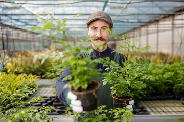 Happy male gardener with mustache offer plants and flowers to the clients in the greenhouse