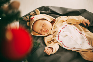 A cute newborn baby boy in a reindeer costume sleeps on a pouf in a room decorated for Christmas and New year. A child sleeps next to a Christmas tree on New year's eve. The concept of Christmas and