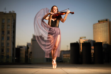 A beautiful adult woman with long hair posing with violin on pointe shoes outdoors in city.