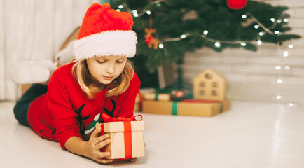 Obraz na płótnie Canvas A blonde girl lies on the floor next to a decorated Christmas tree and holds a gift tied with a red ribbon. The girl unwraps the gift. Christmas and new year concept