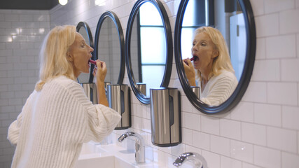 Attractive aged woman paints lips with lipstick in bathroom of restaurant