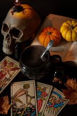 Tea in a cauldron mug with tarot cards, a skull with candle, book, little pumpkins, obsidian crystals and autumn leaves on a black table