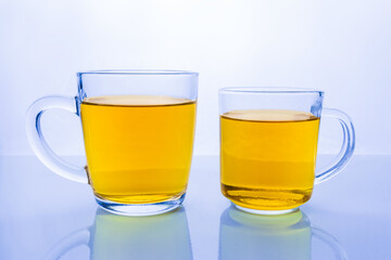 Two transparent glass cups with yellow liquid on reflective surface in blue tint. Mugs of tea or juice