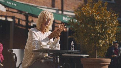 Elegant aged lady disinfecting hands with antiseptic sitting in outdoors cafe