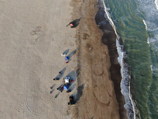 Vista cenital de vehículos y personas descansando sobre la arena, en la costa del océano, durante el atardecer.