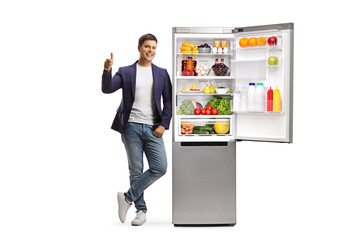 Full length portrait of a young man leaning on a full fridge and showing thumbs up