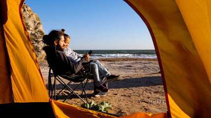  Father sits on camp chair and reads electronic reader for little daughter who sitting on his knees on the beach in windy weather, view from camping tent.