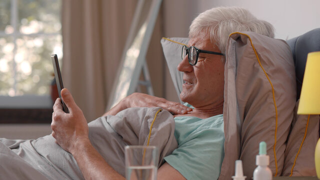Sick senior man holding cellphone and calling doctor online lying in bed