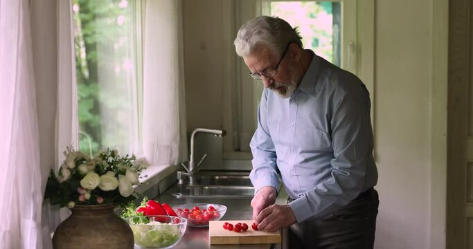 Aged 55s bearded man cutting fresh tomatoes tasting it cooking vegan salad standing alone in kitchen, healthy vegetarian dinner, caring husband prepare surprise for wife, enjoy process, hobby concept