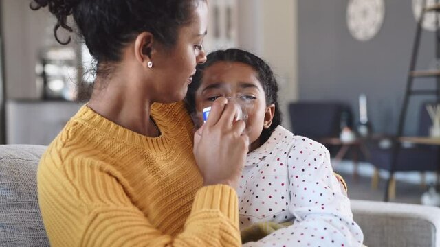 Black mother helping sick daughter use nebulizer while embracing her on couch at home. Woman makes inhalation with equipment to indian girl. Ill child lying on couch having respiratory illness.