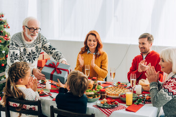 Smiling grandfather giving present to grandson near family applauding at home