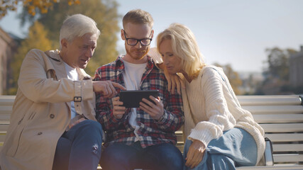 Senior parents and adult son sitting on bench using digital tablet outdoors