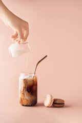 Young female hand pouring milk in glass of iced coffee in tall glass with golden straw. Coffee and macaroons chocolate, vanilla on pastel background for your design. Food concept in vintage style